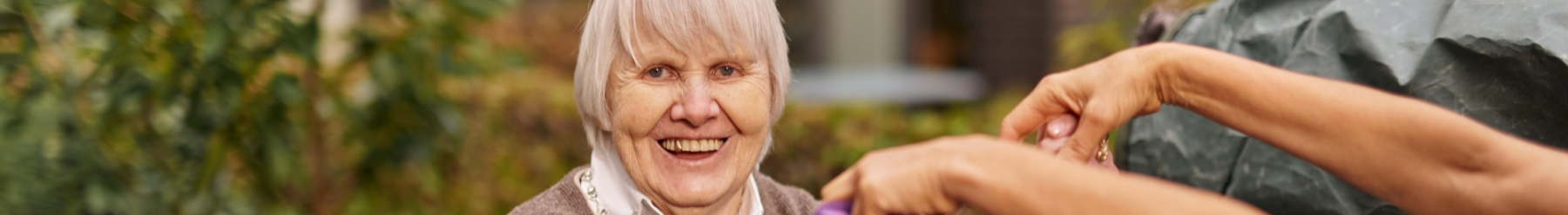 L?chelnde ?ltere Frau mit grauem Haar im Freien, im Hintergrund gr?ne Pflanzen, zwei H?nde halten ihre H?nde wie im Tanz.